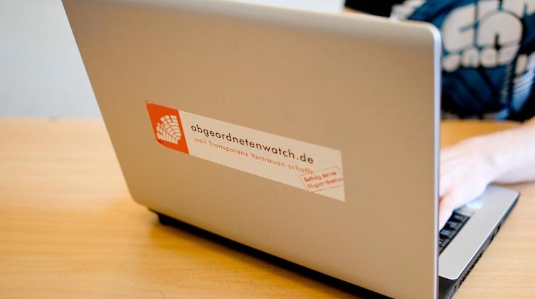Eine Person befindet sich an einem Laptop, auf dem ein Aufkleber mit der Internetseite «abgeordnetenwatch.de» klebt. Foto: Daniel Bockwoldt/Archiv