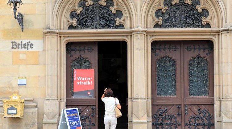 Eine Frau geht auf eine Tür der Hauptpost zu, einer Filiale der Postbank, an der ein rotes Plakat mit der Aufschrift «Heute Warnstreik» hängt. Foto: Peter Gercke/Archivbild