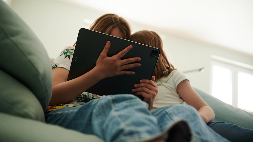 Zwei Schwestern sitzen auf der Couch und gucken in das Tablet ihrer Eltern. / Foto: Annette Riedl/dpa/Symbolbild