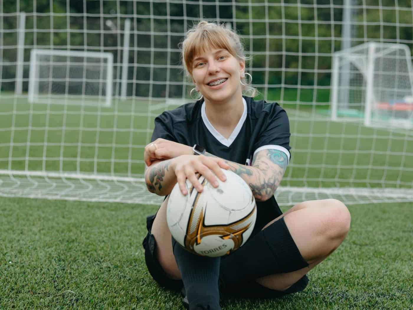 Eine lächelnde Fußballerin hält einen Fußball im Sitzen auf einem Rasenplatz vor einem Tor.