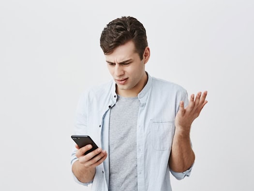 Junger Mann in blauem Hemd schaut verwirrt auf sein Smartphone, die flache Hand fragend erhoben