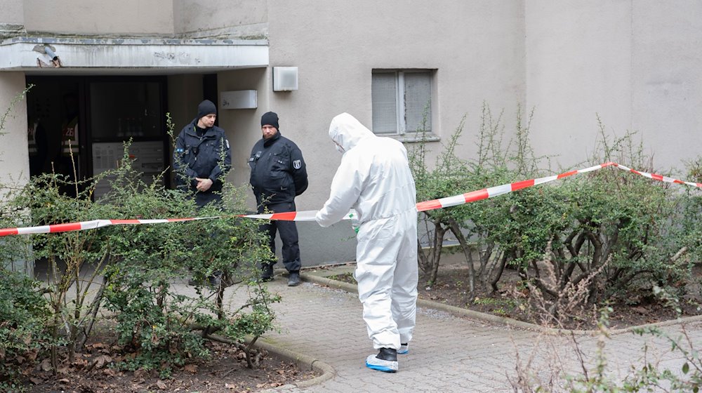 Polizisten stehen am Eingang eines Mehrfamilienhauses in Stadtteil Kreuzberg, während ein Polizist in Schutzanzug an einem Absperrband steht. / Foto: Paul Zinken/dpa