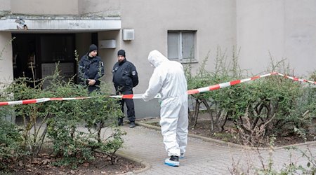 Polizisten stehen am Eingang eines Mehrfamilienhauses in Stadtteil Kreuzberg, während ein Polizist in Schutzanzug an einem Absperrband steht. / Foto: Paul Zinken/dpa