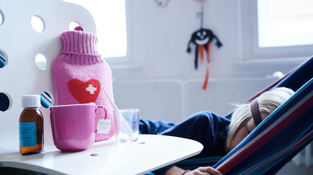 Fieberthermometer, Hustensaft, Wärmflasche und eine Tasse Tee stehen auf einem Stuhl, im Hintergrund liegt ein krankes Kind. / Foto: Annette Riedl/dpa