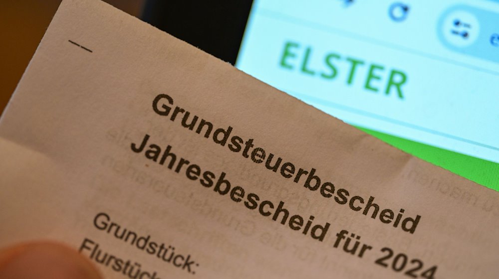 Ein Grundsteuerbescheid für 2024 wird vor einen Computerbildschirm gehalten, auf dem das Logo der Steuerplattform Elster angezeigt wird. / Foto: Bernd Weißbrod/dpa/Archivbild