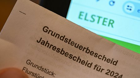 Ein Grundsteuerbescheid für 2024 wird vor einen Computerbildschirm gehalten, auf dem das Logo der Steuerplattform Elster angezeigt wird. / Foto: Bernd Weißbrod/dpa/Archivbild