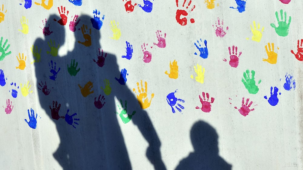 Ein Mann mit einem Kind auf dem Arm und einem an der Hand wirft einen Schatten auf eine mit bunten Handabdrücken bemalte Wand einer Kindertagesstätte. (Symbolbild) / Foto: Peter Kneffel/dpa