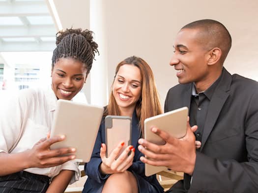 Drei lächelnde Berufstätige in Business-Kleidung betrachten gemeinsam Inhalte auf einem Tablet und einem Smartphone, symbolisieren Teamarbeit und digitale Kommunikation im modernen Arbeitsumfeld.