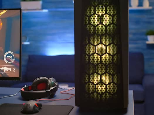Ein moderner Gaming-PC-Arbeitsplatz mit beleuchtetem Gehäuse, großem Monitor, auf dem ein Spiel läuft, sowie Gaming-Headset und Maus auf dem Tisch. Die Szene zeigt die Kombination aus leistungsstarker Hardware und immersivem Entertainment.