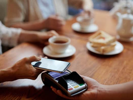 Eine Person bezahlt kontaktlos mit dem Smartphone an einem Kartenterminal in einem Café, während im Hintergrund eine Tasse Tee und ein Teller mit Sandwiches auf dem Tisch stehen.