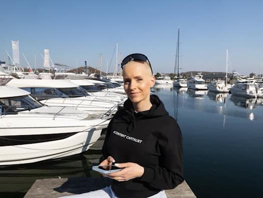 Anike Ekina, Content-Creatorin und Unternehmerin, hält ein Smartphone in der Hand und steht vor einem Yachthafen.