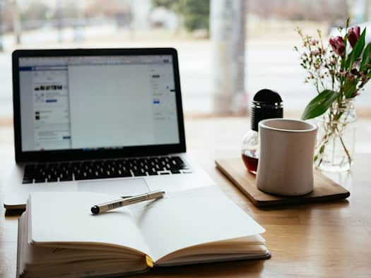 Arbeitsplatz mit offenem Laptop, Notizbuch, Stift, Kaffeetasse und Blumenvasen