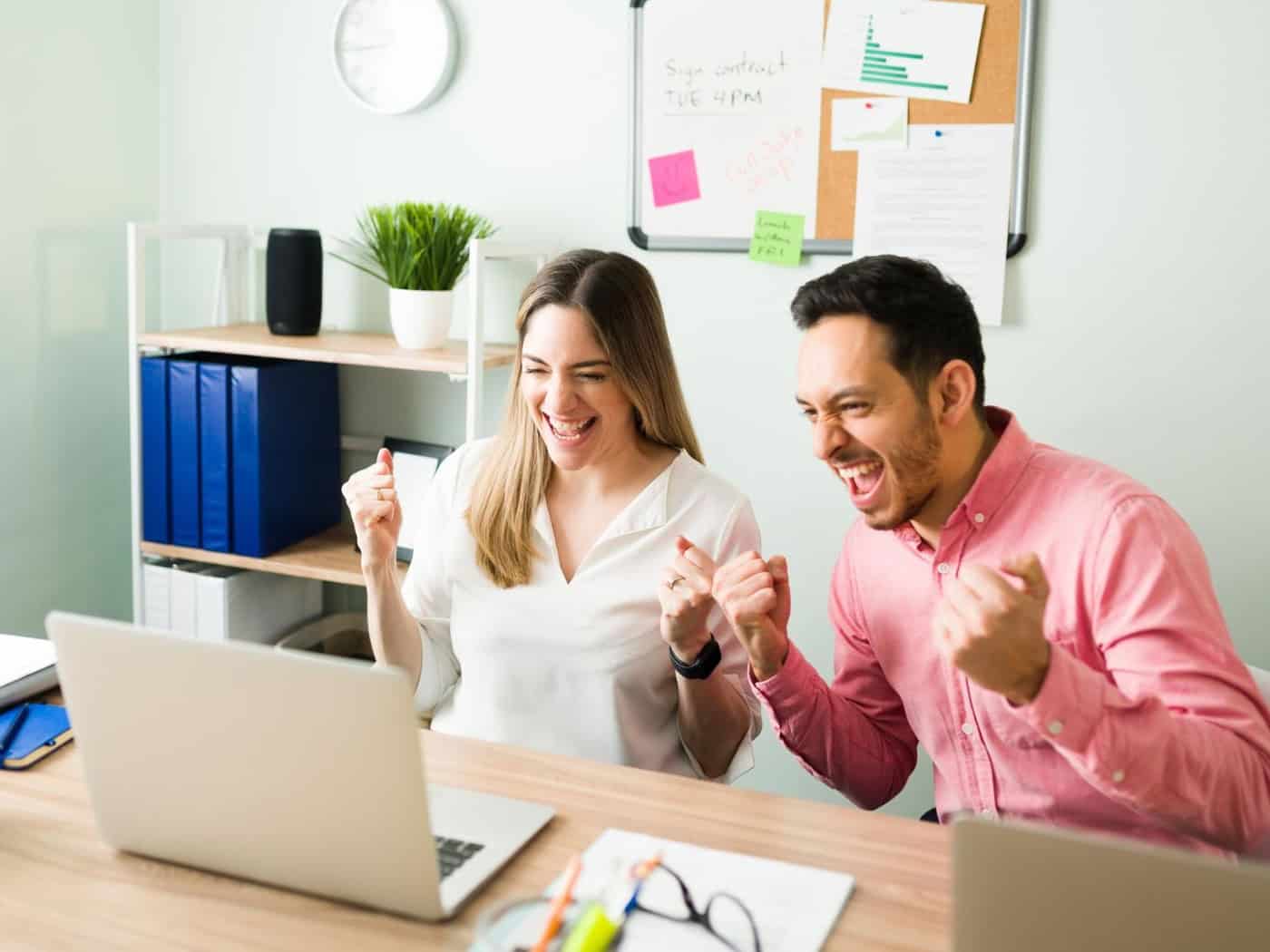 Zwei Personen jubeln vor dem Laptop im Büro.
