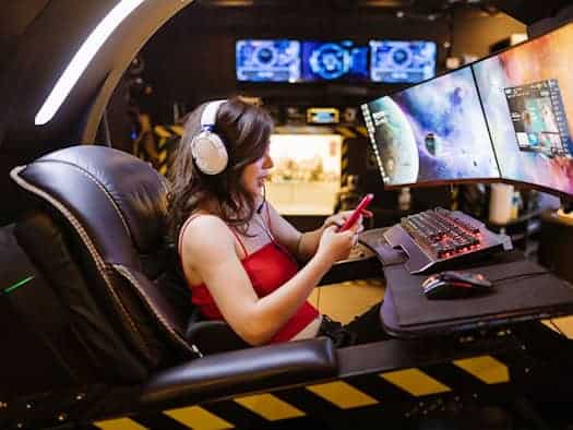 Frau in rotem Kleid spielt mit Smartphone und Headset an einem Gaming-PC mit mehreren Monitoren