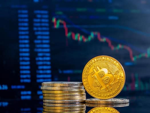 Goldene Bitcoin-Münze auf Stapel kleinerer Münzen, im Hintergrund unscharfer Trading-Chart