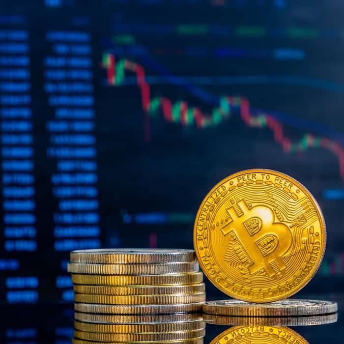 Goldene Bitcoin-Münze auf Stapel kleinerer Münzen, im Hintergrund unscharfer Trading-Chart