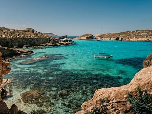 Blue Lagoon auf Comino, Malta: Türkisfarbenes klares Wasser, felsige Klippen und Boote vor blauem Himmel