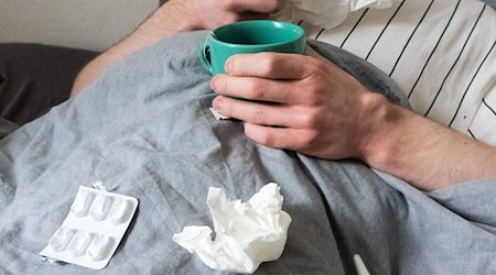 Ein Mann liegt mit Taschentüchern, Teetasse, Nasenspray und Tabletten im Bett. / Foto: Philip Dulian/dpa/Illustration