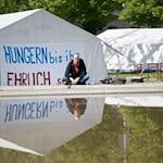 Wolfgang Metzeler-Kick sitzt am neuen Standort des Hungerstreik-Camps des Bündnisses «Hungern bis ihr ehrlich seid» im Regierungsviertel vor einem Banner. / Foto: Sebastian Gollnow/dpa