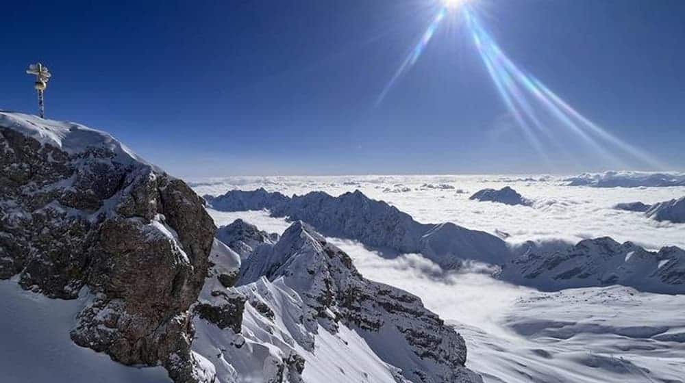 Die Sonne scheint über dem Gipfelkreuz der schneebedeckten Zugspitze. / Foto: Christoph Trost/dpa