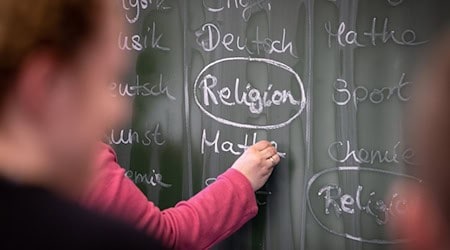 Eine Lehrerin schreibt mit Kreide einen Stundenplan mit Religionsunterricht an eine Tafel. / Foto: Friso Gentsch/dpa