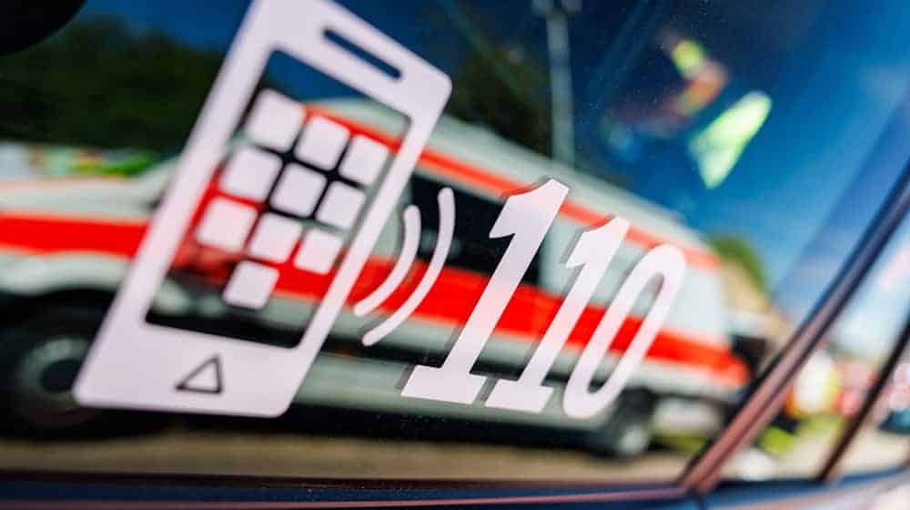 Die Nummer des Polizeinotrufs 110 steht auf der Scheibe eines Polizeifahrzeugs, in der sich ein Fahrzeug vom Roten Kreuz spiegelt. / Foto: Daniel Karmann/dpa