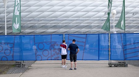 Zwei Männer fotografieren über einen Zaun hinweg die München Fußball Arena. Am 14.06.2024 findet in der Arena das Eröffnungsspiel der Fußball-Europameisterschaft statt. Die Fußball-EM findet vom 14. Juni bis 14. Juli statt. / Foto: Sven Hoppe/dpa