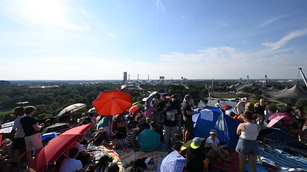 Taylor-Swift-Fans sitzen mit Sonnenschirmen vor der Kulisse des Olympiastadions auf dem Gipfel des Olympiabergs. / Foto: Felix Hörhager/dpa