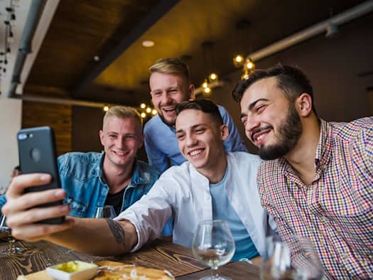 Vier lachende Männer machen ein Selfie in einer Bar, im Hintergrund stehen Weingläser und ein gemütlicher Tisch mit Snacks.