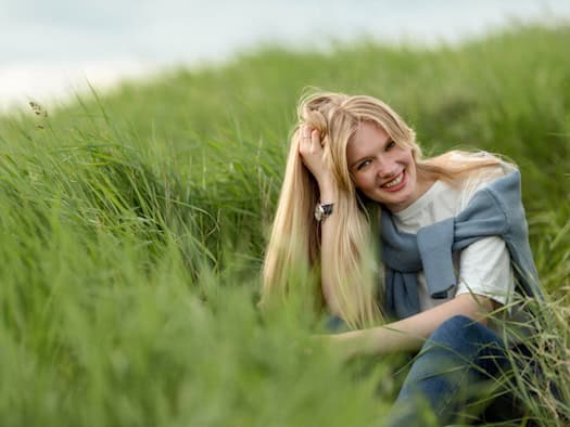 Eine lächelnde Frau mit langem blondem Haar sitzt entspannt in hohem Gras auf einer Wiese – Symbol für die Herausforderungen der Pollensaison.