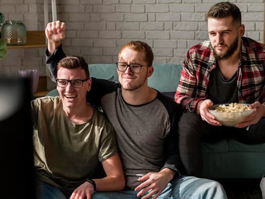 Drei Männer schauen gemeinsam Fernsehen, einer mit Popcorn, gemütliches Wohnzimmer.