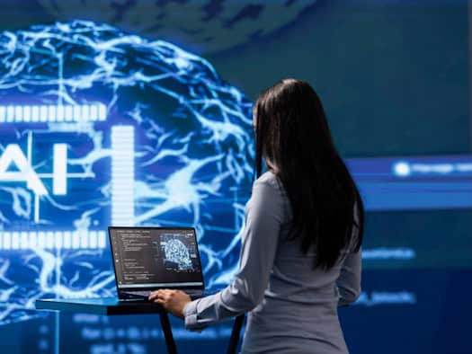 Frau arbeitet an einem Laptop vor einer futuristischen KI-Visualisierung mit Gehirnnetzwerken und dem Wort "AI".