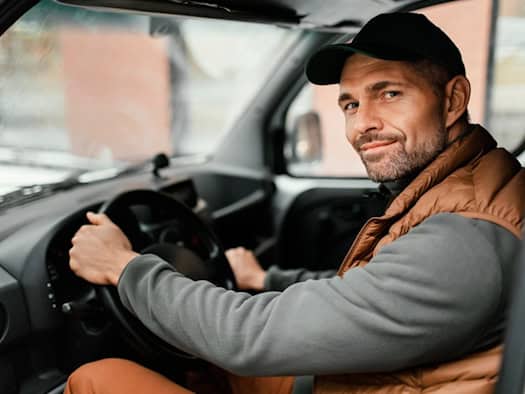 Mann mit Basecap und brauner Weste sitzt lächelnd am Steuer eines Autos und hält das Lenkrad mit beiden Händen.