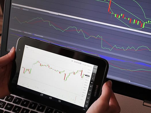 Tablet mit einer Trading-App, die Kursverläufe und Charts anzeigt. Laptop mit Finanzdaten im Hintergrund.