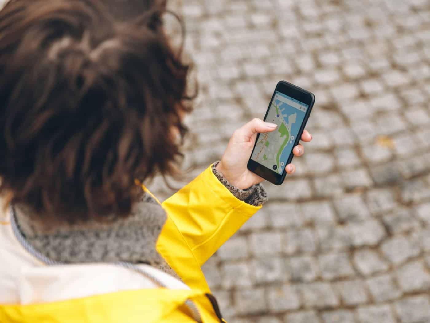 Eine Person in gelber Jacke nutzt ein Smartphone mit einer Navigations-App auf einem gepflasterten Weg.