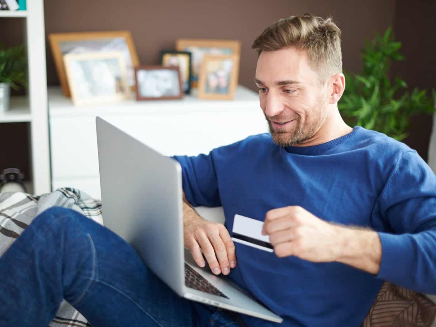 Mann sitzt mit Laptop auf dem Sofa und hält eine Kreditkarte in der Hand, um eine Online-Zahlung durchzuführen