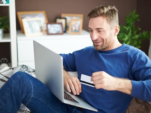 Mann sitzt mit Laptop auf dem Sofa und hält eine Kreditkarte in der Hand, um eine Online-Zahlung durchzuführen