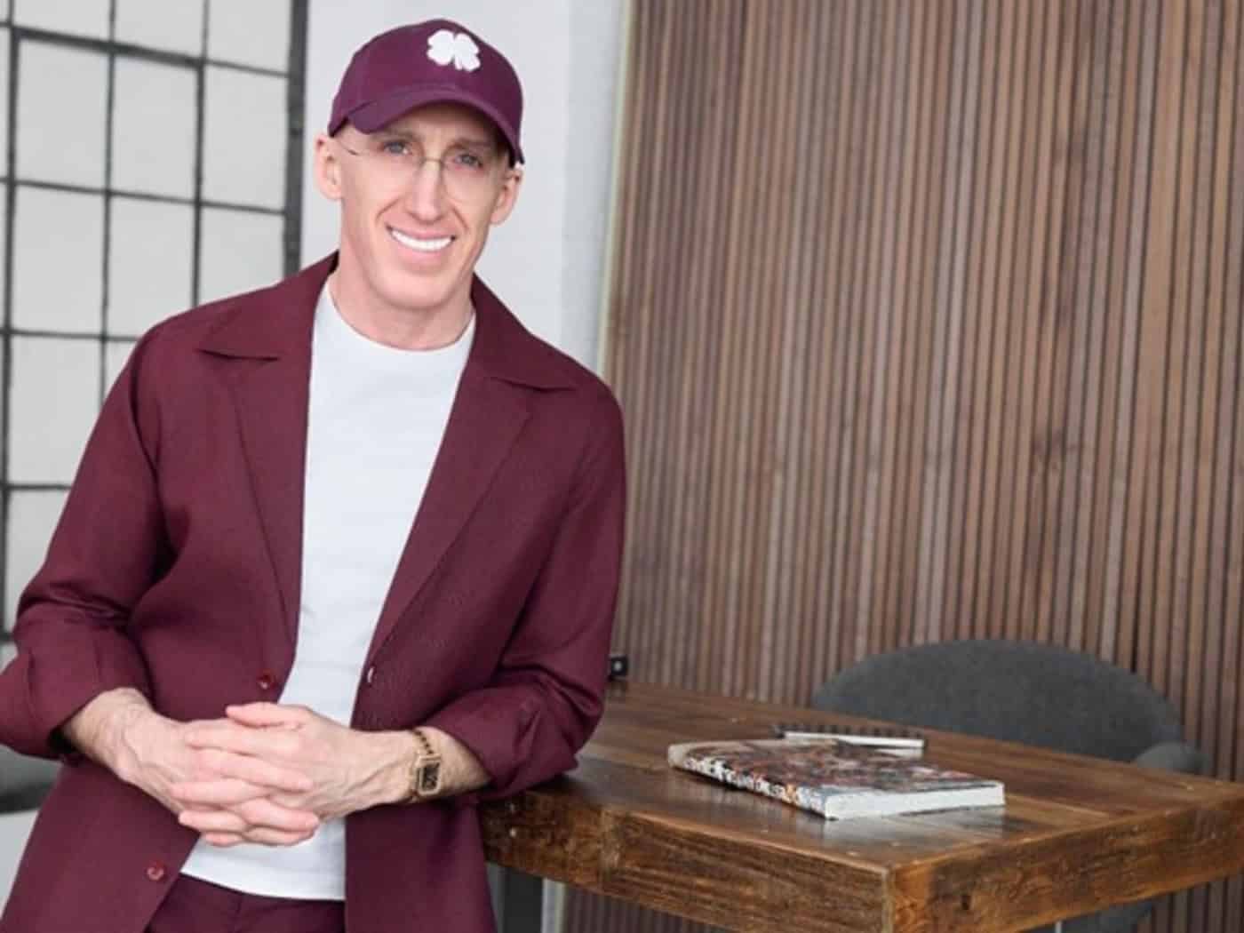 Uri Poliavich lächelnd in weinroter Jacke und Basecap an einem Holztisch