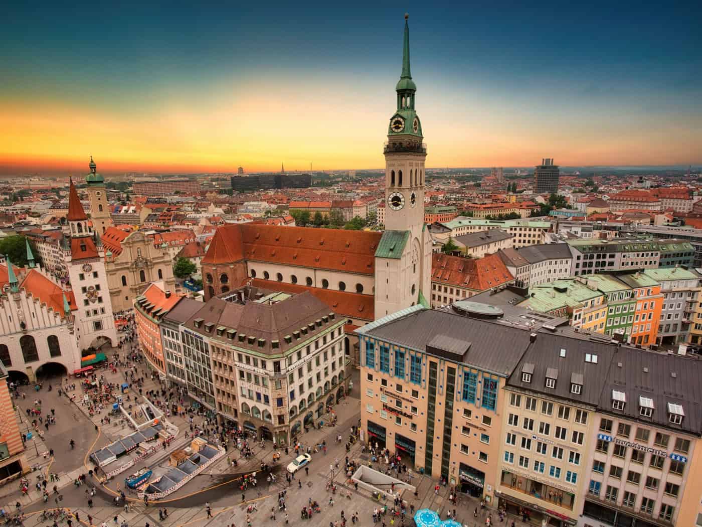 Stadtansicht von München bei Sonnenuntergang mit belebtem Platz, historischen Gebäuden und einem Kirchturm