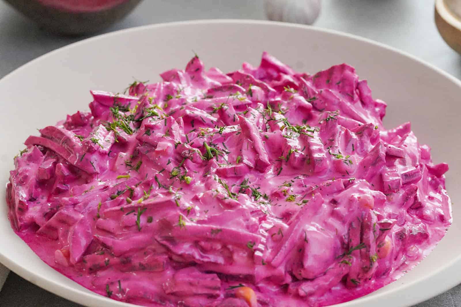 In einem weißen, tiefen Porzellanteller ist ein lila Rote-Beete-Salat angerichtet. Dahinter liegt Knoblauch und eine rote Zwiebel.