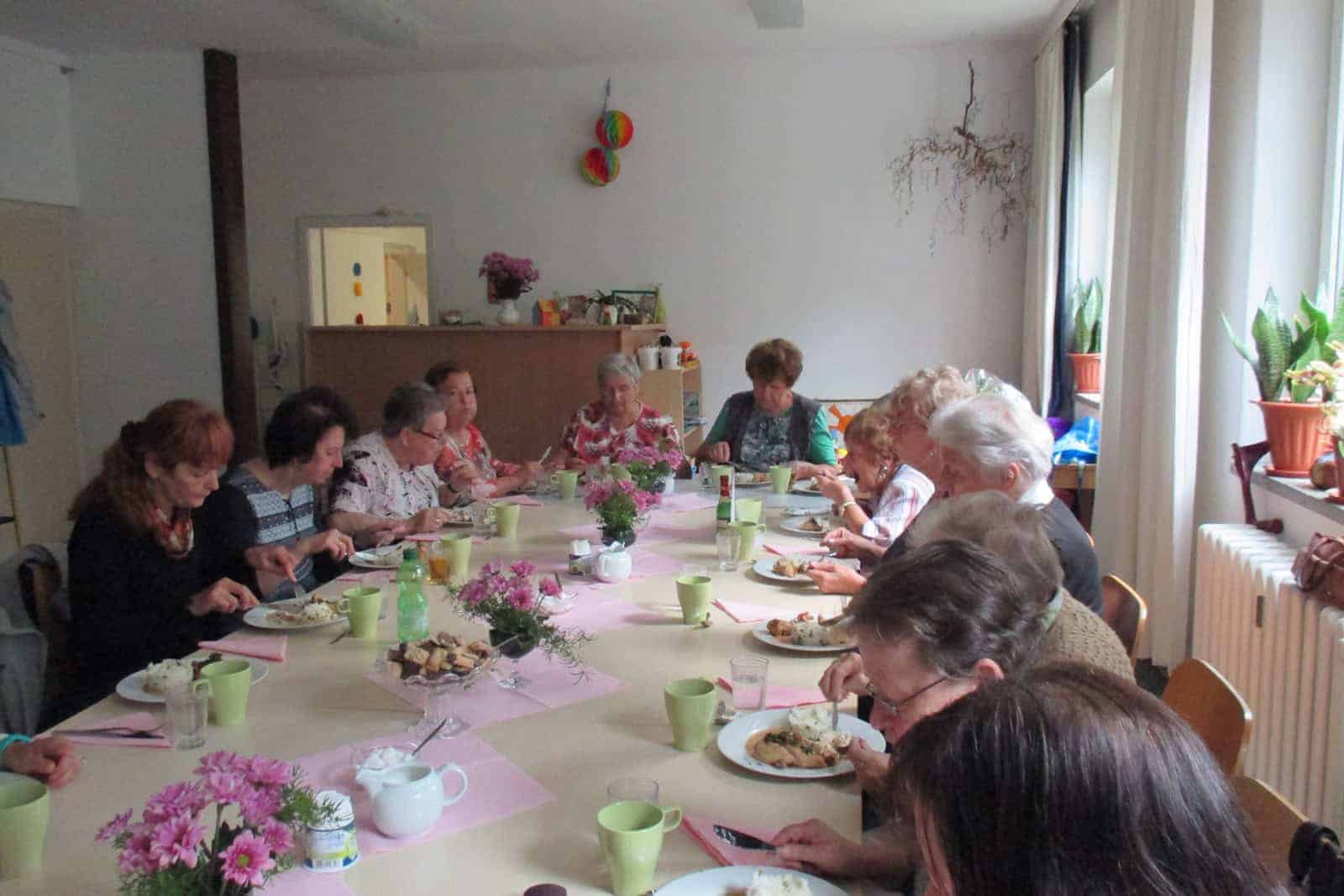Eine Gruppe älterer Menschen sitzt an einer Tafel, isst und kommuniziert. In der Mitte stehen kleine Blumengestecke. Foto: MGH Bernsdorf