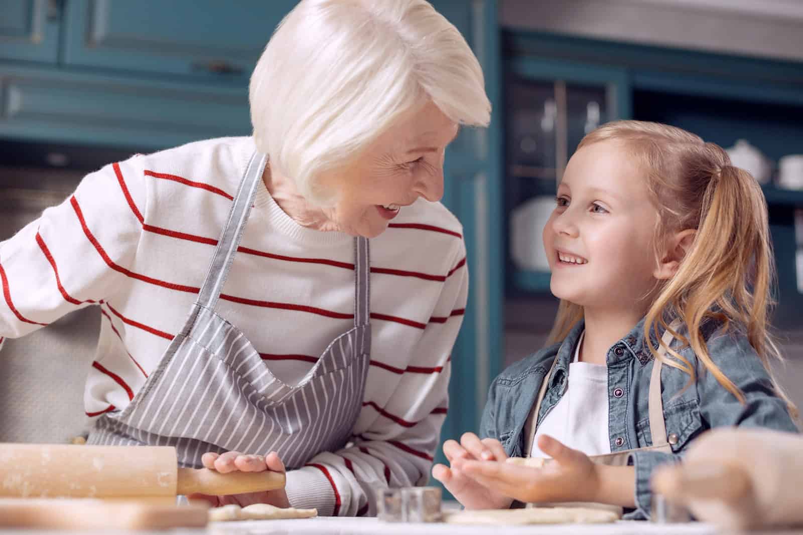 Eine weißhaarige Seniorin mit Schürze und ein lächelndes Mädchen mit Zöpfen rollen gemeinsam den Teig aus. Foto: AdobeStock/zinkevych