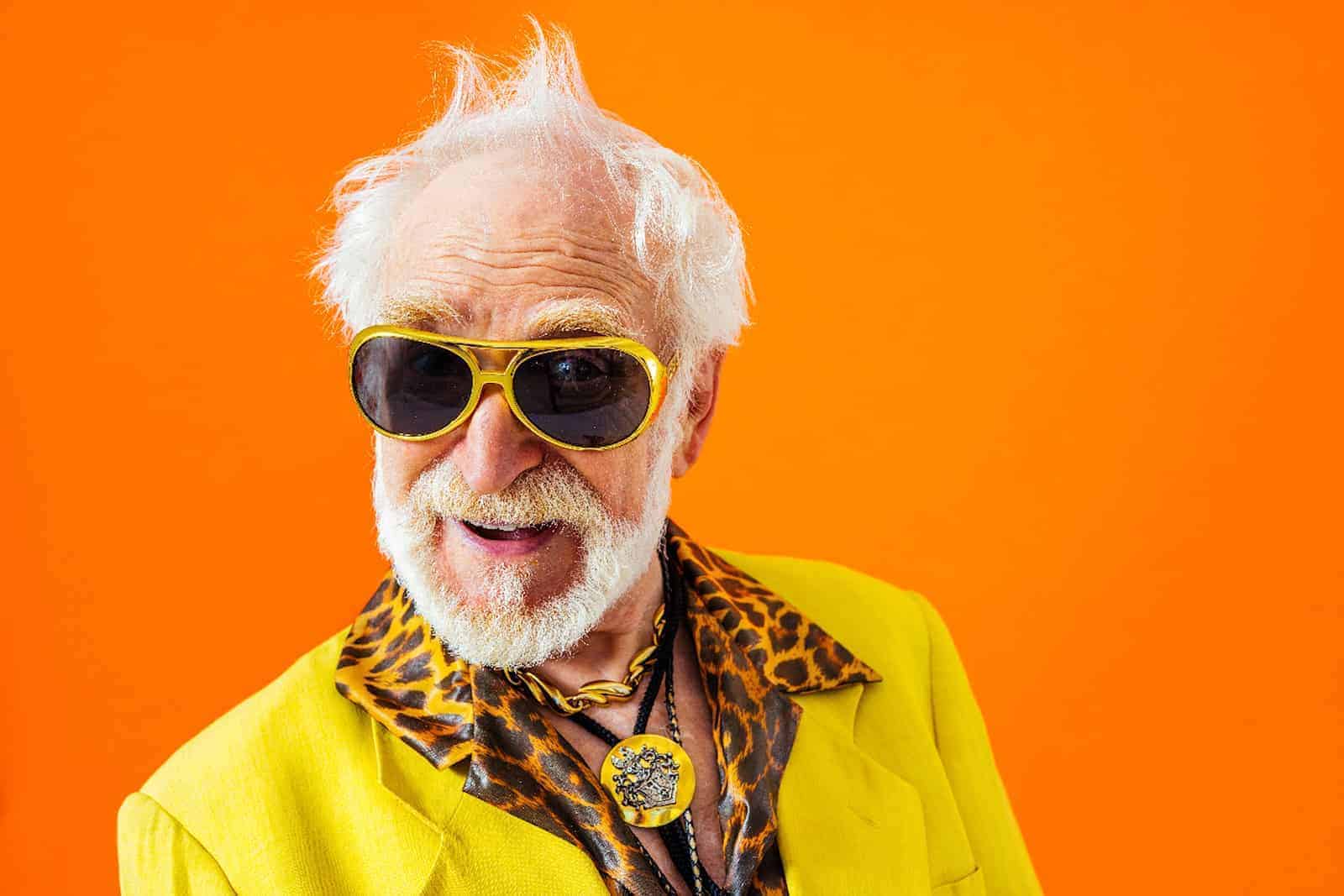 Modebewußter älterer Herr mit knallig bunter Anzugjacke und Sonnenbrille und buntem Hintergrund.