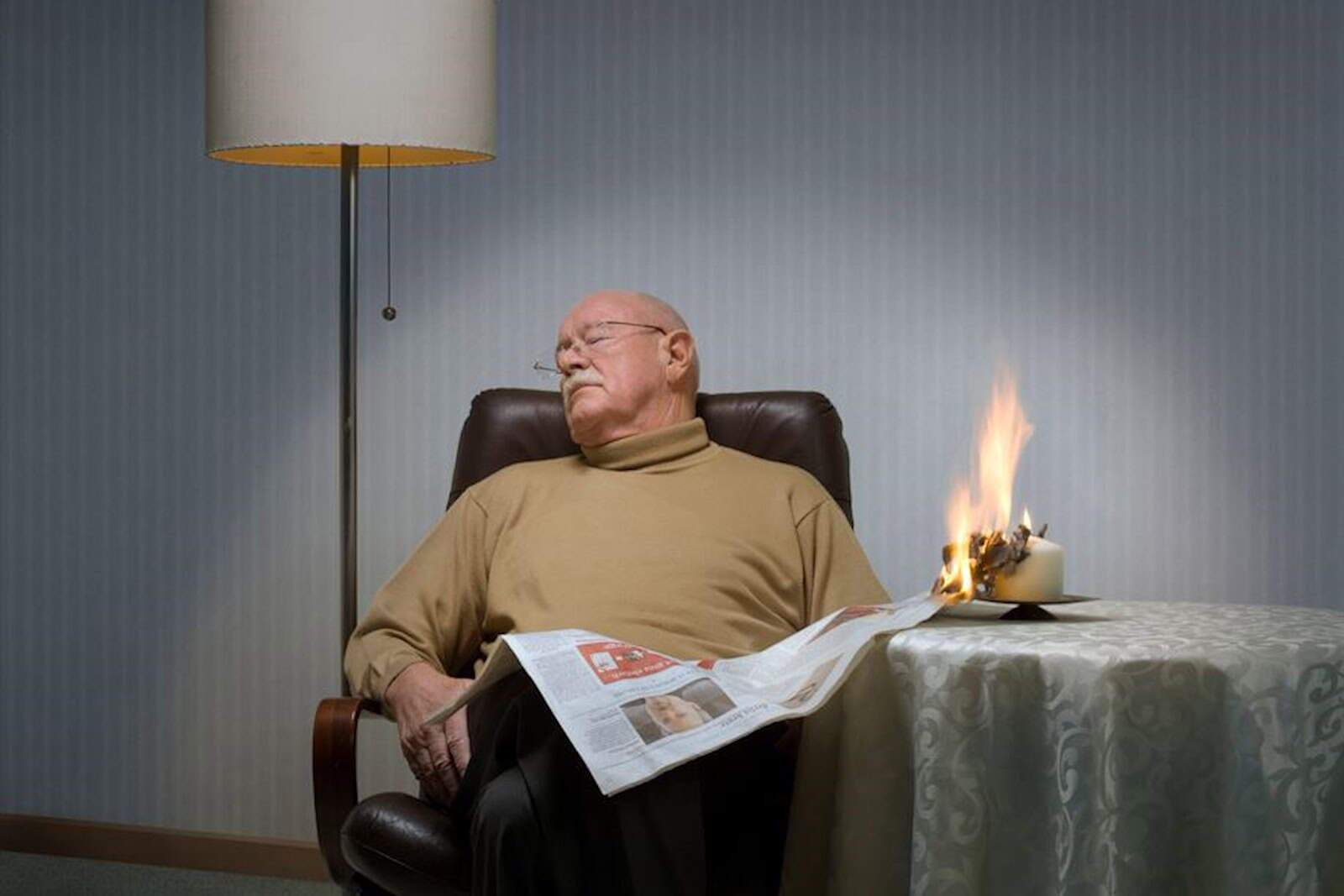 Ein Senior hält eine Zeitung in der Hand und ist eingeschlafen. Das Papier hat an einer Kerze Feuer gefangen. Foto: Forum Brandrauchprävention