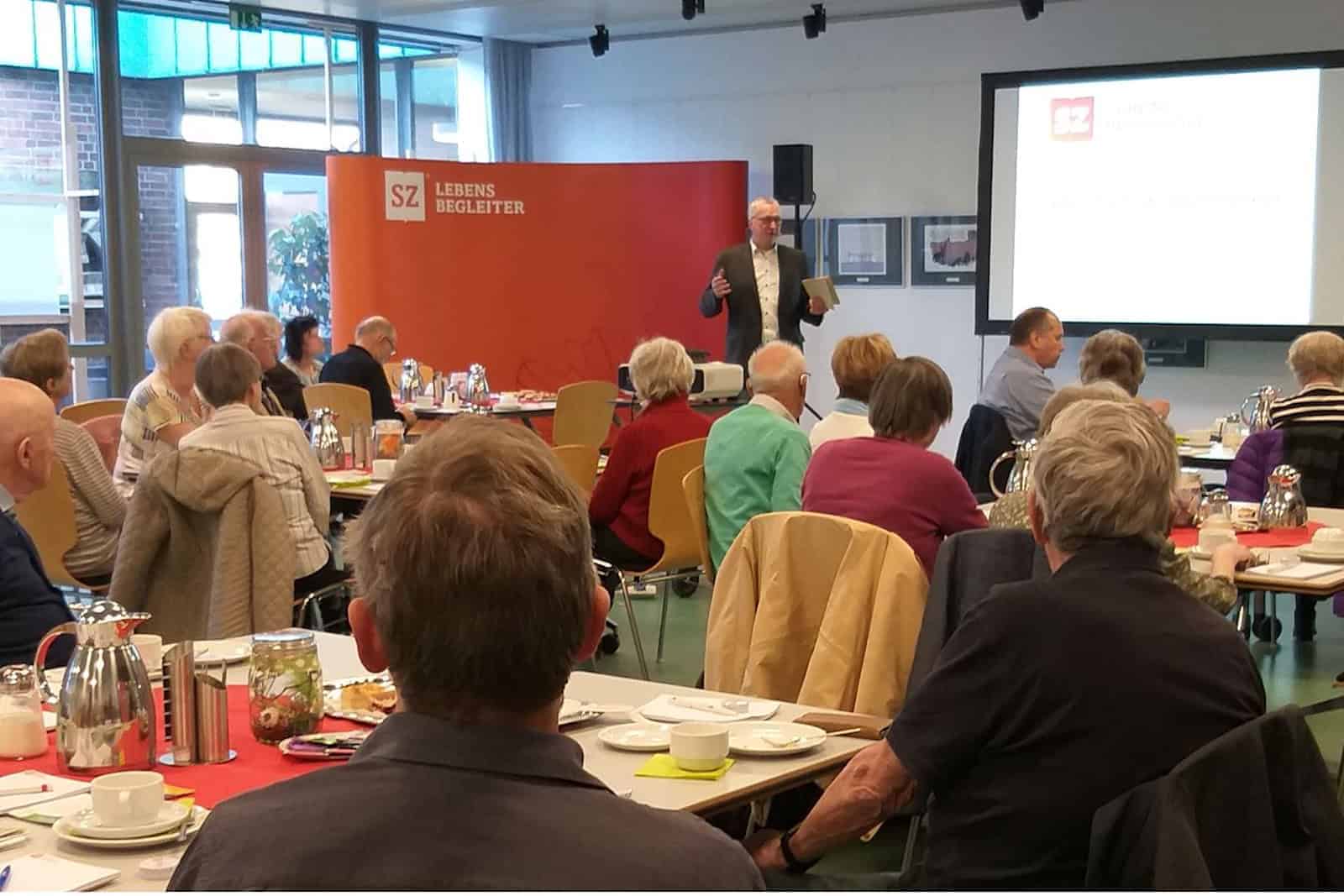 Im Veranstaltungssaal sitzen viele Gäste bei Kaffee und Kuchen und hören dem Moderator vom Gesundheitsvortrag zu.