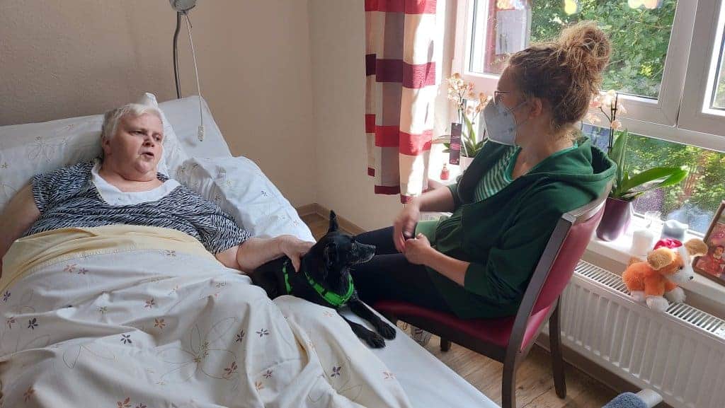 Lena Scheidig mit Hund Tarana zu Besuch im Pflegeheim bei einer Seniorin. Hund liegt auf Bett.