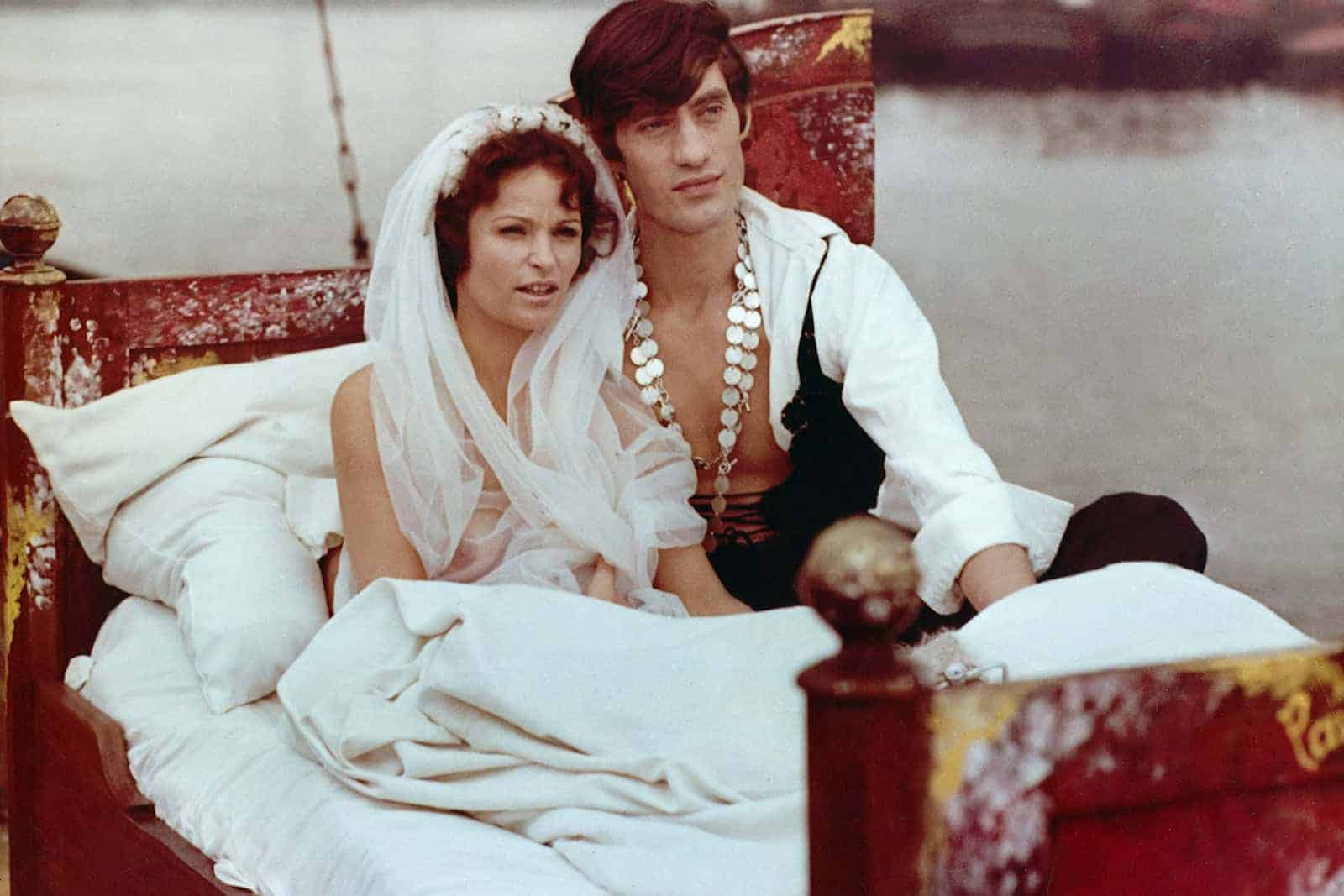 Die Schauspielerin Angelica Domröse ist mit einem Blumenkranz geschmückt. Sie sitzt gemeinsam mit ihrem Paul in einem Bett, dass auf einem Spree-Kahn steht. Foto: DEFA-Stiftung/Herbert Kroiss, Manfred Damm
