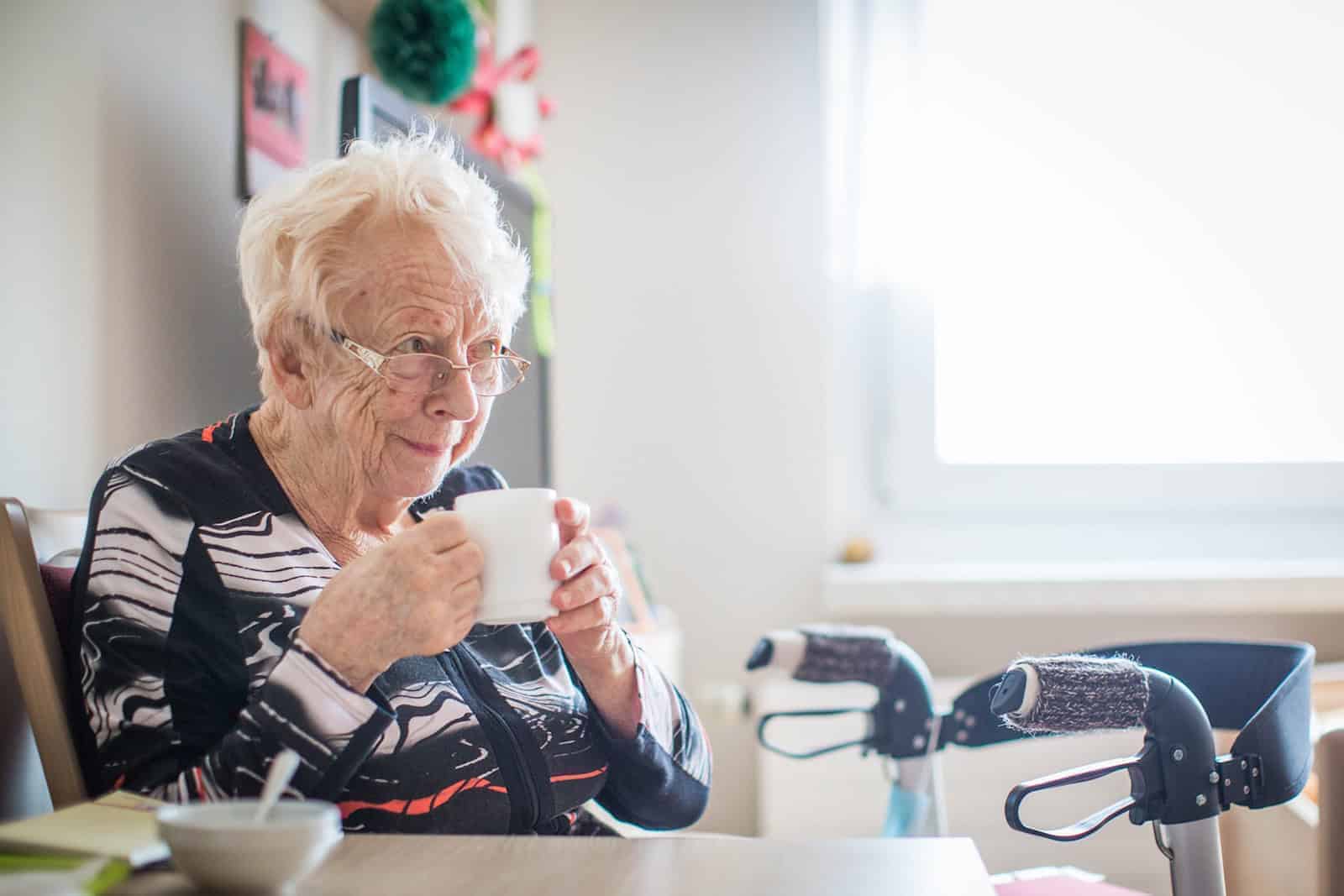 Seniorin Rosemarie John lächelt zufrieden mit Kaffee in der Hand am Tisch im Seniorenheim.