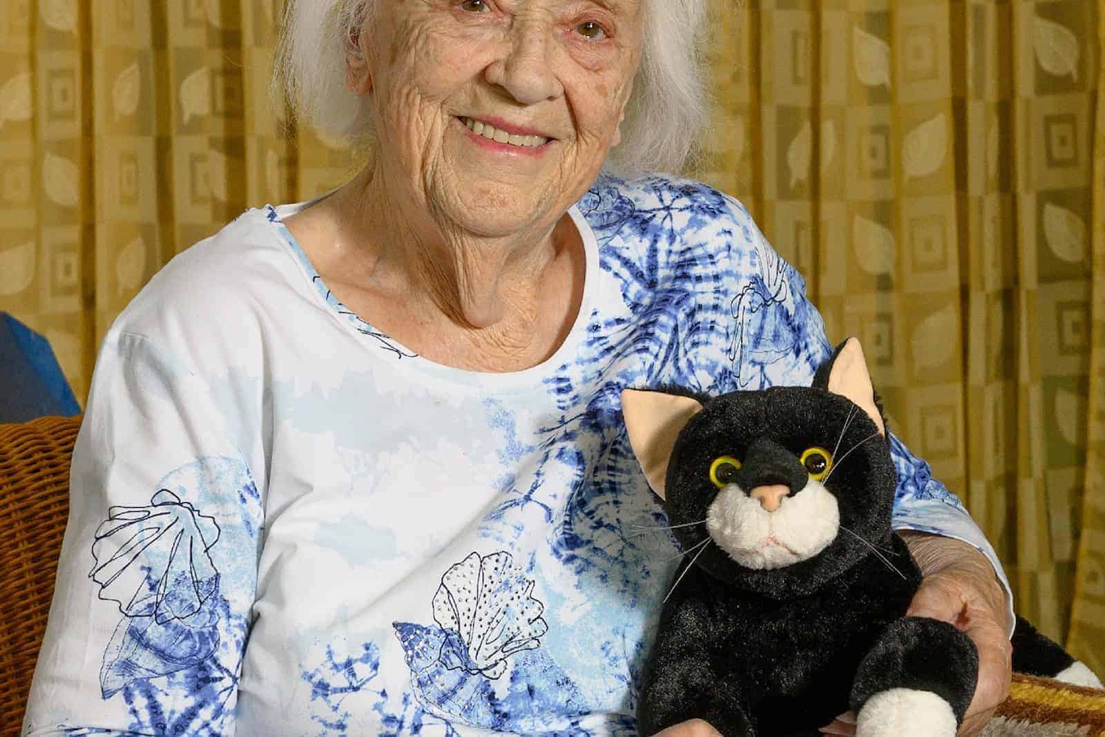 Eine Seniorin sitzt im Sessel und streichelt glücklich eine Stoffkatze.