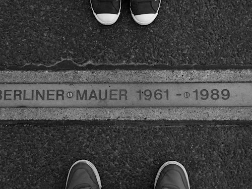 Schwarz-Weiß-Foto mit der Bodenmarkierung „Berliner Mauer 1961–1989“. Auf beiden Seiten stehen Menschen, von denen nur die Schuhe zu sehen sind.